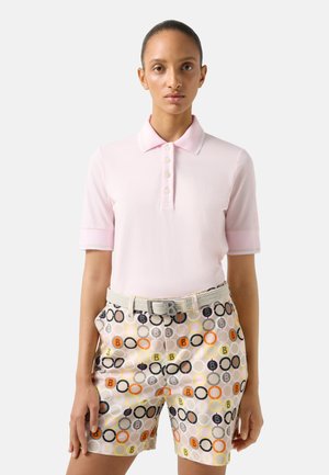 Ženský model má na sobě světle růžové polo tričko s krátkými rukávy a krátké kalhoty s vysokým pasem s vícebarevným kruhovým vzorem a tkaným opaskem.