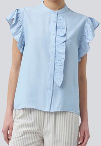 Blusa sin mangas de color azul claro con volantes en el frente y botones, combinada con pantalones blancos con delgadas rayas verticales grises.