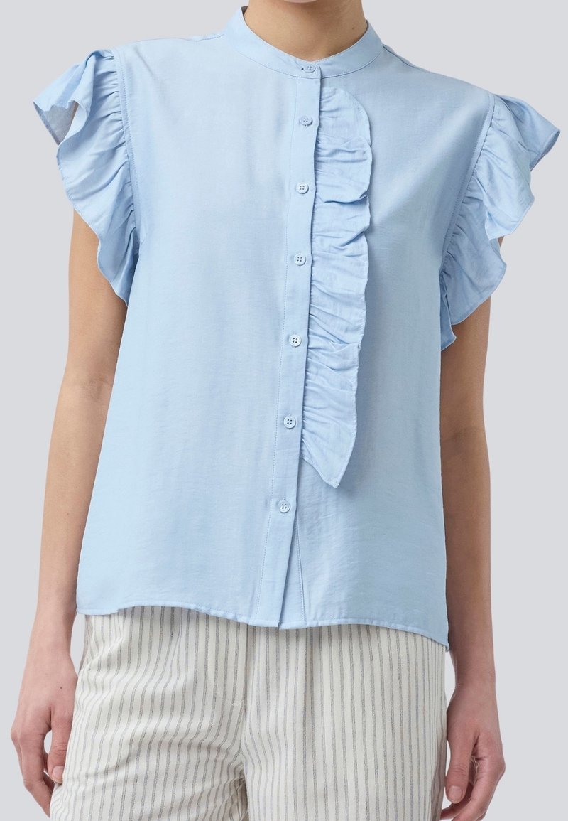 Blusa sin mangas de color azul claro con volantes en el frente y botones, combinada con pantalones blancos con delgadas rayas verticales grises.