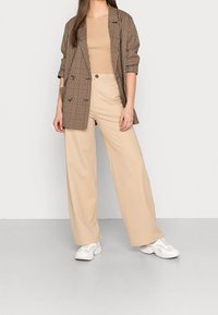 Femme portant un pantalon large beige, un top à côtes beige, un blazer à carreaux marron avec des manches retroussées et des baskets blanches, se tenant devant un mur uni.