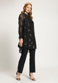 Zwarte doorschijnende tuniek met een textuurpatroon, gecombineerd met zwarte broeken. Inclusief zwarte handtas en hoge sandalen. Elegante en verlengde silhouette.