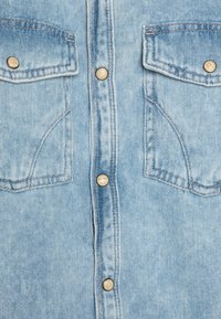 Camicia in denim azzurro chiaro con chiusura a bottoni, due tasche sul petto con pattina e bottoni automatici color oro. Tessuto texturizzato con cuciture visibili.