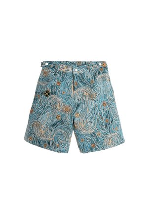 Joven de pie, vistiendo una camiseta gráfica beige con siluetas y texto, pantalones cortos azules con estampado, calcetines blancos y botas marrones, fondo blanco liso.