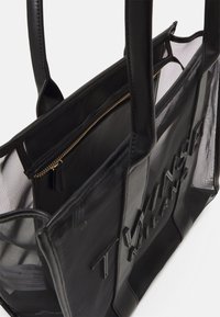 Sac cabas transparent noir avec poignées en cuir, pochette intérieure zippée visible et texte embossé "Twinset Milano" sur le panneau avant.