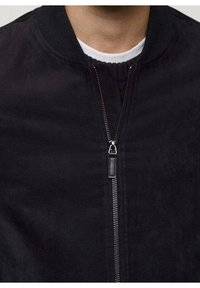 Mann, der eine schwarze Zip-Jacke über einem weißen Rundhals-Shirt trägt, mit Fokus auf dem Reißverschluss und dem Kragenbereich der Jacke.