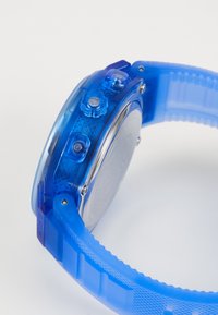 La montre bleue transparente présente un cadran rond, trois boutons latéraux, un bracelet en silicone texturé et un dessous métallique avec un détail de marque.