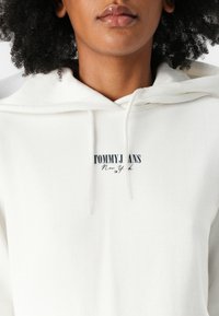 Witte hoodie van zachte stof, met een buidelzak en trekkoorden. "TOMMY JEANS New York" logo in zwart en rood op de voorkant.