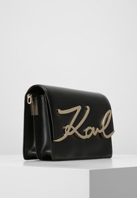 Černá kožená kabelka s strukturovaným designem, na přední straně výrazný zlatý podpis „Karl“ a kovový detail na zipu.