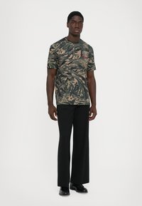 Joven de pie vistiendo una camiseta de tigre en verde y beige, pantalones anchos negros y zapatos de vestir negros, contra un fondo blanco.
