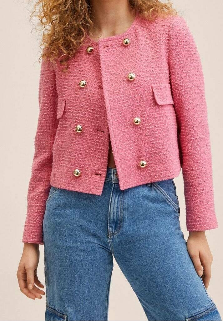 Femme portant une veste rose texturée avec des boutons dorés et un jean bleu taille haute, debout, les bras détendus le long du corps.