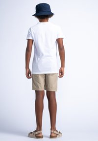 Witte katoenen t-shirt, beige shorts en een marineblauwe bucket hat. Casual schoenen met een gestructureerde zool en verstelbare banden. Achteraanzicht.