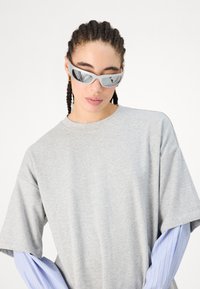 Szürke, oversized póló kerek nyakkal; világoskék és fehér csíkos hosszú ujjúak láthatók. Szürke napszemüveg, tükröződő lencsékkel.