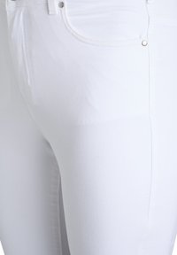 Jeans elásticos blancos con una textura suave, con un diseño de cinco bolsillos, acentos de hardware metálico y una silueta ajustada.