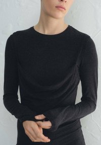 Černý top s dlouhým rukávem z měkké a elastické látky, s volným výstřihem do tvaru lodi a manžetami s otvory na palce; hladká textura s detailem volného záhybu vpředu.