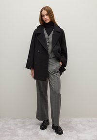 Schwarzer zweireihiger Mantel über einer grauen Weste und weit geschnittenen Hosen. Der Stoff wirkt weich, mit Knopfdetails und glänzenden schwarzen Stiefeln.