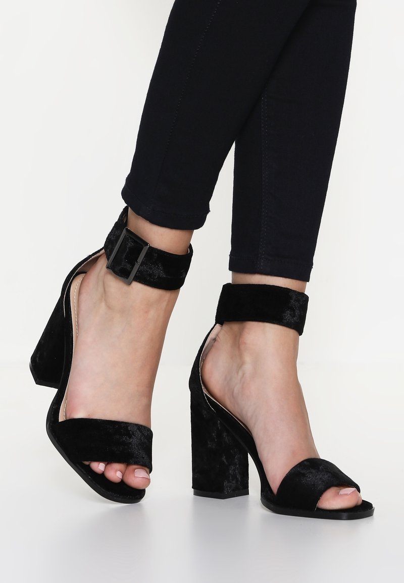 Pieds chaussés de sandales à talons blocs en velours noir avec brides à la cheville, associées à un pantalon noir ajusté sur fond blanc.