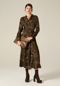 Abito avvolgente marrone con motivi circolari, maniche lunghe, vita legata, abbinato a una pochette lucida marrone e sandali neri con tacco a blocco.