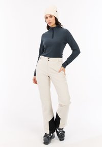 Donna che indossa un berretto a maglia crema, una maglietta a maniche lunghe con zip blu navy, pantaloni da neve beige e stivali da sci neri, in piedi su uno sfondo bianco.