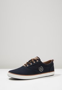 Marinblå avslappnad sneakers med bruna snören, vit sula och broderad logotyp på sidan, visad på vit yta mot grå bakgrund.