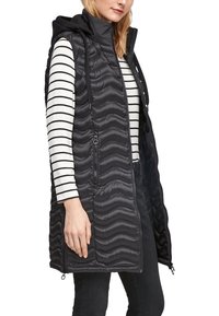 Schwarze gepolsterte Weste mit Kapuze, ausgestattet mit wellenförmiger Steppung, seitlichen Reißverschlusstaschen und einem durchgehenden Frontreißverschluss. Getragen über einem gestreiften langärmeligen Oberteil.
