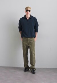 Young man wearing dark sunglasses, navy blue long-sleeve shirt, olive green pants, and black shoes standing against a plain gray wall.
