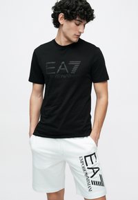 Czarna bawełniana koszulka z tonowanym logo "EA7 Emporio Armani" na przodzie, zestawiona z białymi szortami ze ściągaczem i wyraźnym czarnym logo.