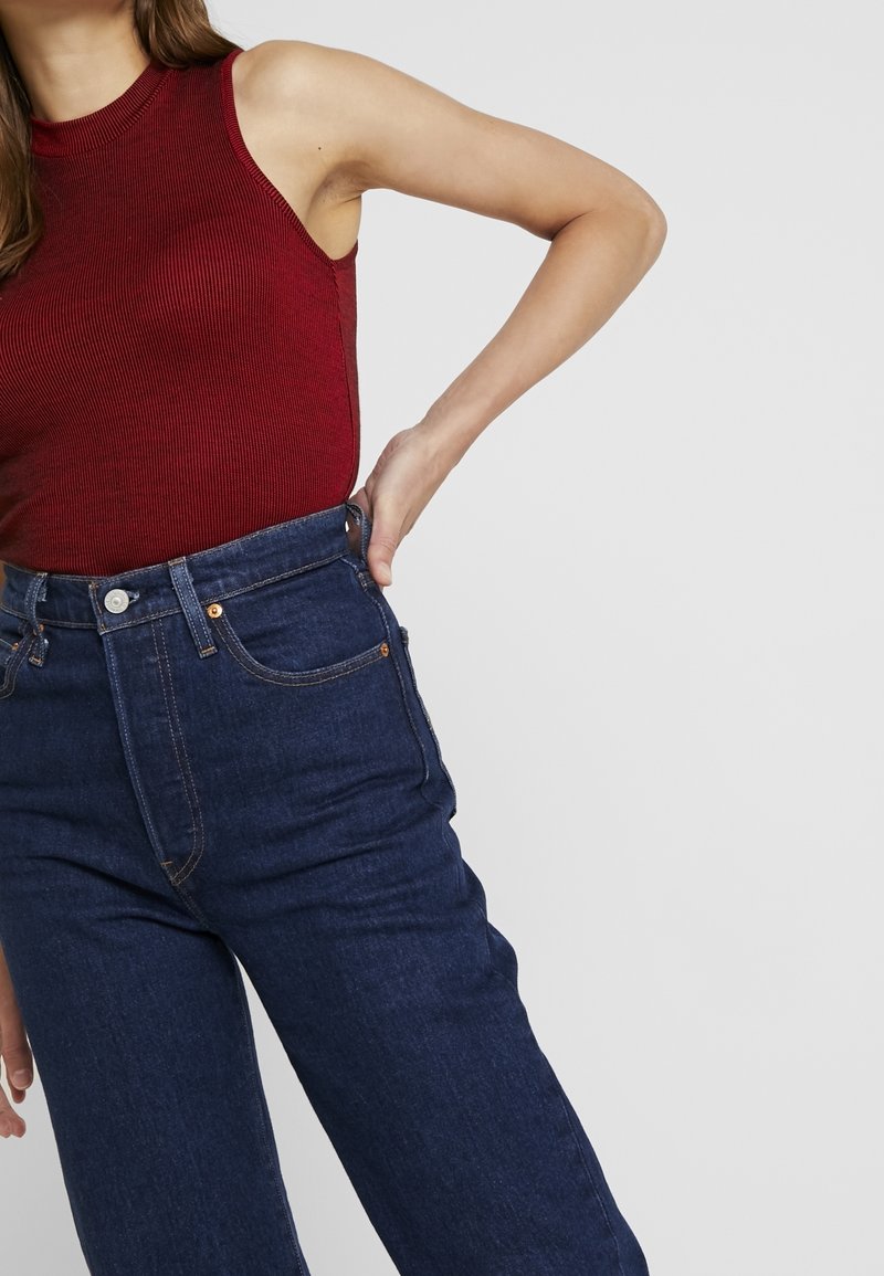Femme portant un haut sans manches rouge côtelé et un jean bleu foncé taille haute, avec une main posée sur la hanche.