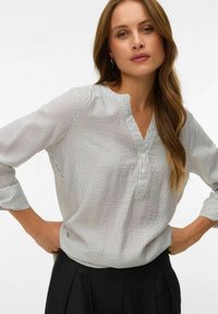 Femme debout, les mains sur les hanches, portant un chemisier blanc à manches longues avec de fines rayures verticales noires et un pantalon noir, fond neutre.