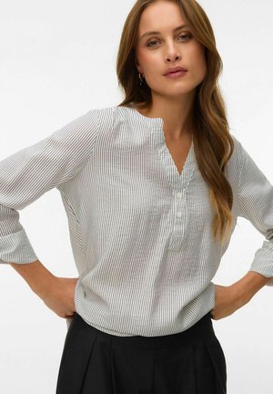 Femme debout, les mains sur les hanches, portant un chemisier blanc à manches longues avec de fines rayures verticales noires et un pantalon noir, fond neutre.