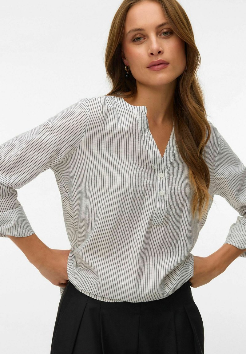 Femme debout, les mains sur les hanches, portant un chemisier blanc à manches longues avec de fines rayures verticales noires et un pantalon noir, fond neutre.