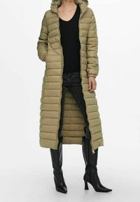 Mujer vistiendo un abrigo acolchado largo de color verde oliva con capucha, parte superior negra, jeans oscuros y botas de tacón alto negras hasta la rodilla de pie.