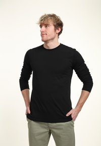 Černé tričko s dlouhým rukávem a kulatým výstřihem vyrobené z hladké látky, má přiléhavé rukávy. Model má na sobě světle zelené kalhoty.