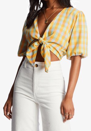 Kobieta ubrana w żółto-niebieską kraciastą krótka bluzkę wiązaną z przodu z bufiastymi rękawami, łączoną z białymi spodniami z wysokim stanem i złotą biżuterią.