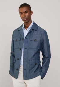 Niebieska kurtka o fakturze z czterema przednimi kieszeniami i guzikami, noszona na jasnoniebieskiej koszuli w paski, z wyrafinowanym krojem i kołnierzykiem.