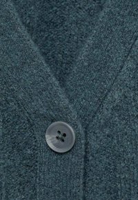 Temno teal volneni kardigan z teksturirano površino, ki ima sivo gumb s štirimi luknjami. Gumbi so nameščeni blizu ovratnika v obliki črke V.