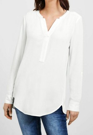 Femme portant un chemisier blanc à manches longues avec un col en V et un jean en denim bleu, debout devant un fond clair uni.