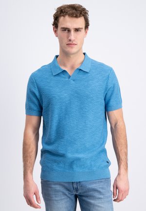 Młody mężczyzna o kręconych włosach, ubrany w teksturowaną jasnoniebieską koszulkę polo z krótkim rękawem i jasnoniebieskie dżinsy, stojący na tle jednolitego białego tła.