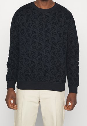 Sweat-shirt bleu marine foncé à manches longues avec un motif floral texturé, porté avec un pantalon beige clair, montrant le torse et les bras.