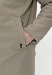Ljus beige ytterjacka med en sidficka, slät textur och en märkesetikett. Ärmen har en ren och avslutad manschett.