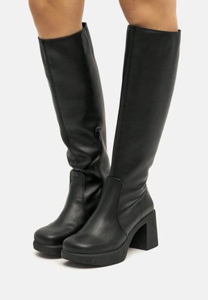 Bottes hautes en cuir noir avec talons blocs et fermetures éclair latérales portées sur des jambes nues sur fond blanc.