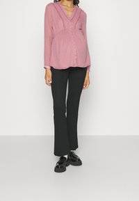 Chemisier rose clair à manches longues avec des pois texturés, encolure en V, boutons sur le devant et taille froncée, assorti à un pantalon noir évasé et des chaussures noires.