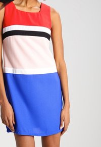 Šaty bez rukávů s červeným vrškem, bílými a černými horizontálními pruhy, přecházejícími do modré sukně. Hladká látka se jednoduchým áčkovým střihem.