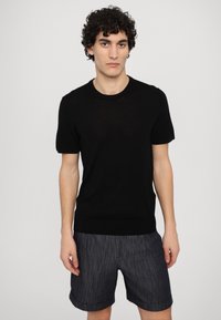 Black short-sleeve knitted sweater with a round neckline and a fitted hem, paired with grey patterned shorts. Smooth texture.