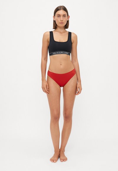 Black sports bra with a wide white logo band, paired with high-cut red bikini briefs. Smooth fabric with a fitted design.