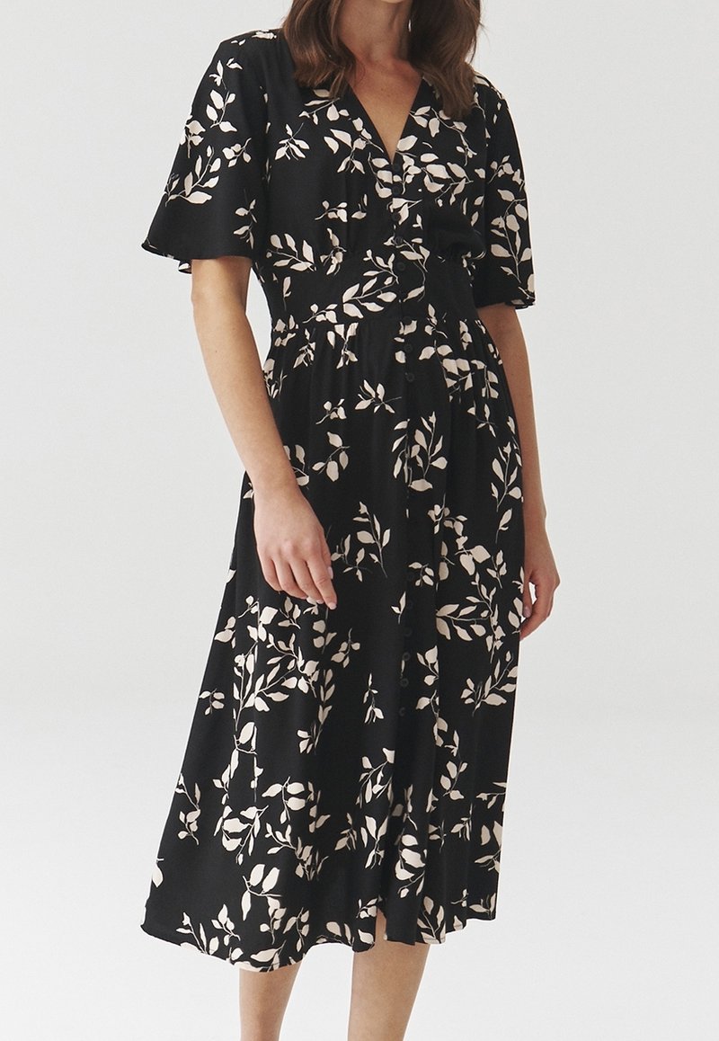 Femme portant une robe midi noire à manches courtes, col en V, avec un imprimé feuilles couleur crème, debout devant un fond clair uni.