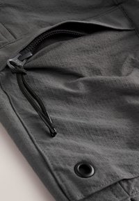 Grijze stof met een gestructureerd patroon, voorzien van een zwart ritsvak en verstelbare trekkoord voor een veilige sluiting. Detail van metalen klinknagel.