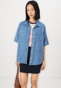 Cămașă din denim de culoare albastru deschis, cu un model texturat, mâneci scurte, două buzunare pe piept și nasturi argintii, asortată cu o fustă bleumarin și o geantă maro.