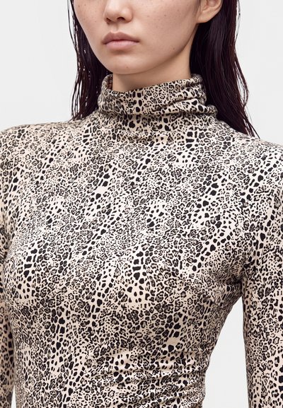 Femme portant un top à col roulé à manches longues beige et noir à imprimé léopard, visible des épaules au menton, avec des cheveux bruns mouillés.