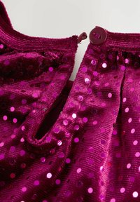 Fuchsia paljettyg med en mjuk, texturerad yta. Har rund halsringning, knäppning framtill med knappar och en delvis öppen dragkedja.