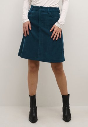 Vrouw die een knie-lange corduroy rok in teal draagt, een wit shirt met lange mouwen en zwarte enkellaarsjes, staand tegen een effen achtergrond.